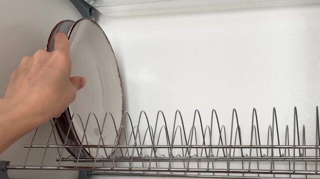 Woman stacking stylish beige plates in a dish rack inside a kitchen cabinet close-up.