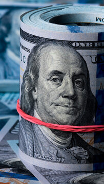 Close-up of rolled of hundred dollar bills with a rubber band around it lying on a pile of cash