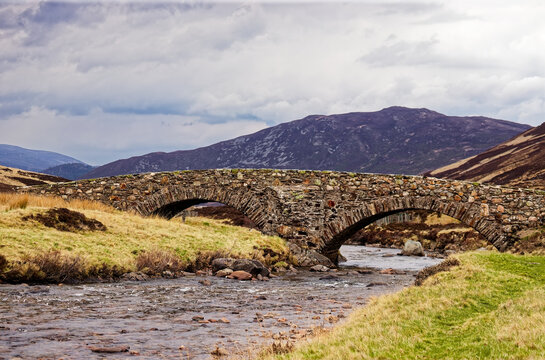 A picturesque view of a traditional historic stone packhorse bridge spanning a flowing river. Set against a backdrop of rugged mountains and a moody, overcast sky in a wild highland meadow. This image