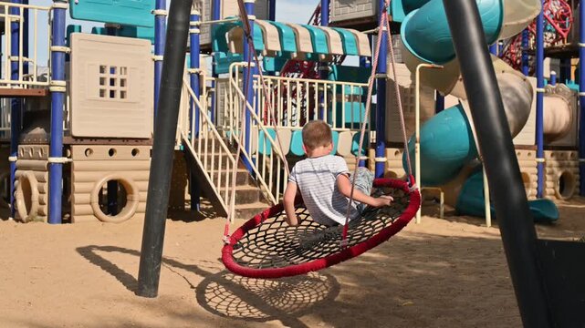 A preschool boy plays on a playground in the summer, swinging on a swing.