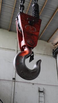 A heavy-duty orange crane hook with a 5-ton capacity slowly lowers from a warehouse ceiling via a thick metal chain, ready for industrial lifting operations in a workshop setting.