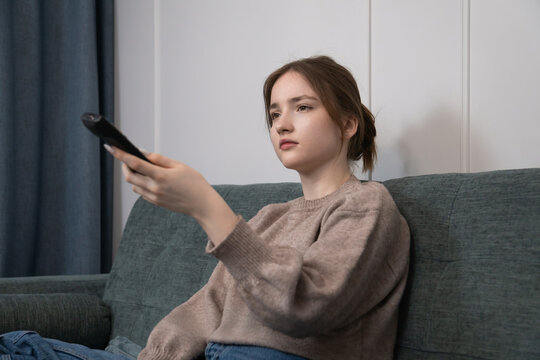 Girl sitting on a comfortable sofa in a living room, holding a tv remote control, watching television and looking bored or unamused while spending leisure time at home