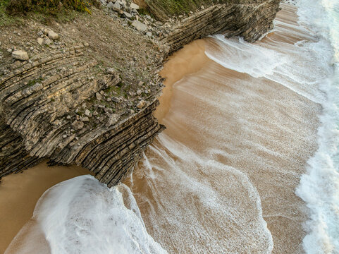 Aerial view of waves crashing against the layered cliffs and sandy shore, creating a dynamic interplay of textures and tones, Buarcos-Figueira da Foz, Figueira da foz, Portugal.