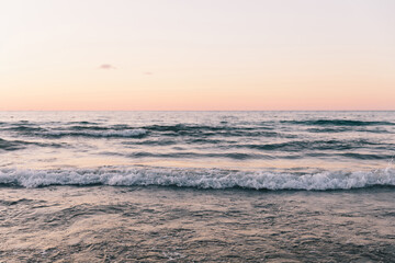 Fototapeta premium Calm ocean waves with white foam roll towards the shore under a soft pink and peach twilight sky. The minimalist seascape captures a tranquil and meditative atmosphere during the golden hour.