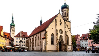 Obraz premium A European square featuring a large church with a tall steeple, surrounded by colorful buildings and trees. People are present