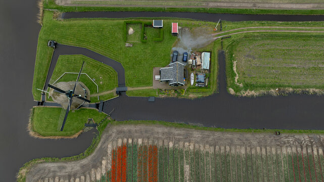Aerial view of a serene windmill standing guard beside a cozy farmhouse, embraced by canals and vibrant tulip fields, 't Zand, Noord-Holland, Netherlands.