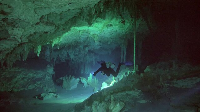 A diver shines a torch across a large underwater cavern revealing rock formations and suspended particles in Cenote Dos Ojos, Mexico
