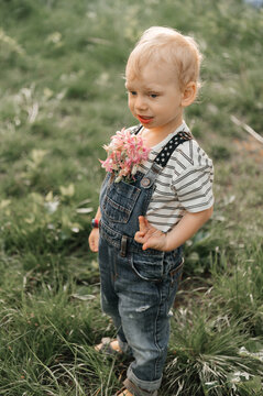A charming little boy with blonde hair, dressed in stylish denim overalls and a straw hat, enjoys a sunny day in a blooming garden. He holds dandelions and spring flowers, 
