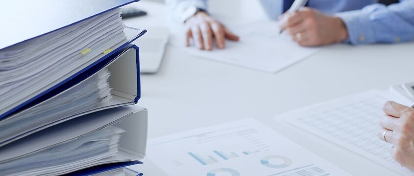 Group of accountants are analyzing financial reports and making notes while sitting at a white table, close up of a stack of blue folders full of paperwork. Audit and taxes in business