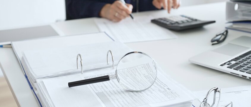Magnifying glass and calculator are on the table over financial documents opposite female accountant analyzing data, reviewing reports, binders and laptop on a white desk. Audit and taxes concept
