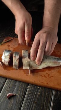 Hands slicing Clupea fish fillets on wooden cutting board with garlic and spices scattered on dark wooden surface in kitchen setting