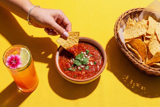 hand dipping a mexican corn chip or tortilla chip in a spicy salsa dip, top view of a bowl of tortillas with salsa on Yellow slate background

