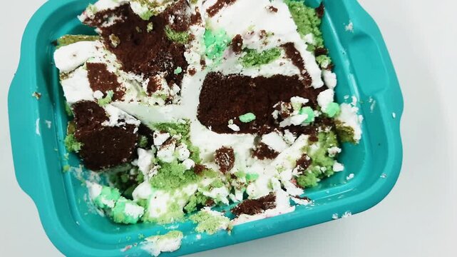 Messy remnants of chocolate and green mint cake with white frosting are rapidly melting inside a bright turquoise plastic container viewed from above motion.