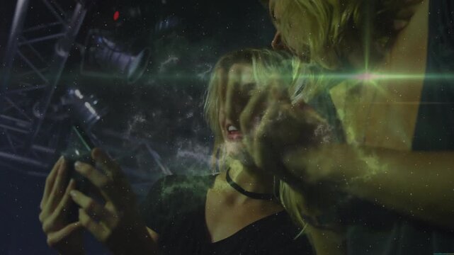 Two women raising phone, leaning in, sharing post, laughing onstage while green mist overlaying