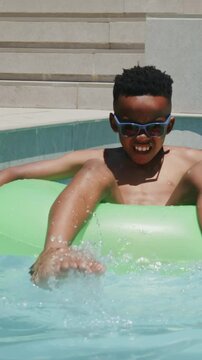 Vertical video: Kicking boy in trunks on sunny day, splashing water at pool in green tube, shades