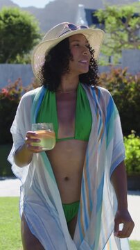 Vertical video: Sun drawing gaze, woman in green bikini walking with glass, turning, savoring drink