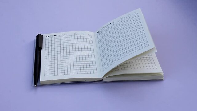 An open notebook with a grid pattern and a black pen lies beside it. Background is a soft purple color, creating a calm workspace atmosphere