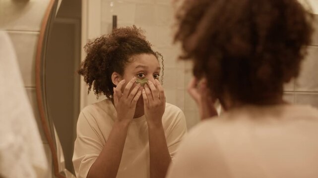 Curly haired African American woman looking in mirror while applying eyepatches during morning routine