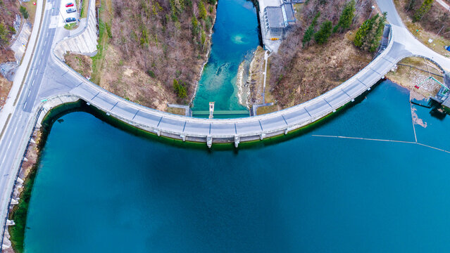 Aerial view of a concrete dam curving against the deep blue water, contrasting with the pale road and the lush green trees, Klaus an der Pyhrnbahn, Oberosterreich, Austria.