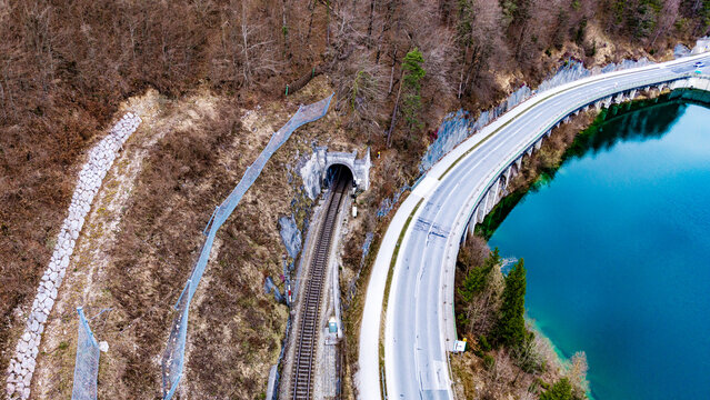Aerial view of a train track leading into a dark tunnel next to a long bridge over the vibrant turquoise lake water, Klaus an der Pyhrnbahn, Oberosterreich, Austria.