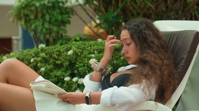 Young woman editing manuscript on island lounge chair marking pages with pen concentrated expression tropical hedge backdrop novelist vibe sunlit leisure creative flow footage for writing and literary