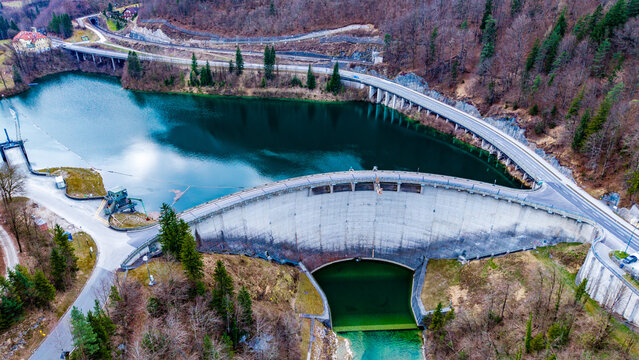 Aerial view of the imposing dam structure reflecting a deep blue, embraced by lush green trees and curving roads, Staudamm Klaus, Klaus an der Pyhrnbahn, Oberosterreich, Austria.