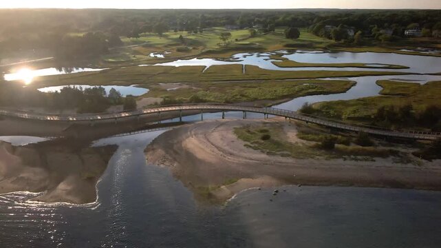 Stunning drone footage captures a curved pedestrian bridge crossing tidal marshlands and estuaries during sunset, with golf course visible in background.