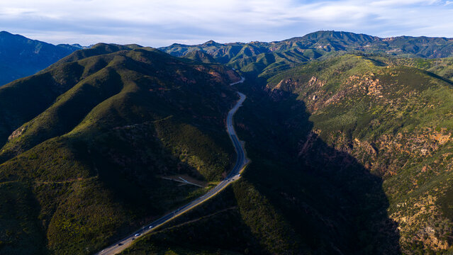 Aerial view of a serpentine road winding through the verdant, rugged mountains, where shadows dance and peaks touch the sky, Malibu, California, United States.