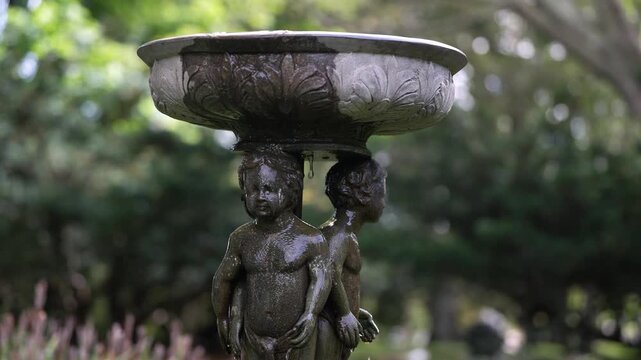 A classical stone fountain featuring cherub figures supporting a decorative basin, set against a lush green garden backdrop with soft bokeh lighting.