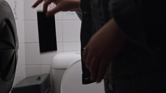 A young woman accidentally drops her phone into a toilet in a dimly lit bathroom, reacting with shock and frustration as it slips from her hand &mdash; close-up