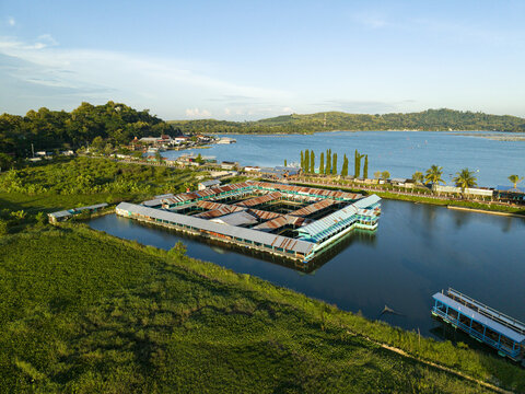 Aerial view of a floating food stall on Rawa Jombor Lake, with emerald water reflecting the sky, contrasting with the red roofs, Klaten, Central Java, Indonesia.