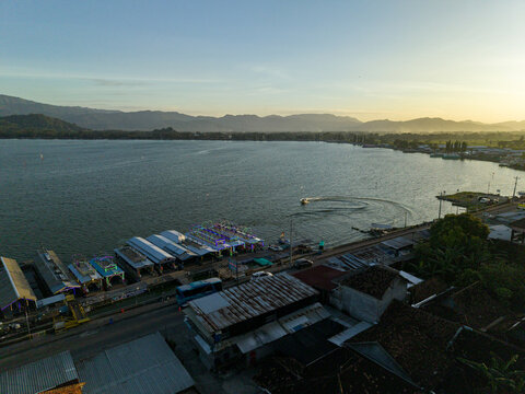 Aerial view of Rawa Jombor lake glistening under the sunlight, with boats docked along the shore, framed by distant mountains, Klaten Regency, Jawa Tengah, Indonesia.