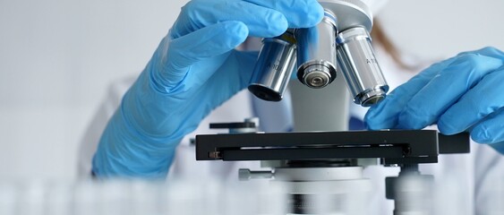 Close up of scientist's gloved hands adjusting microscope, conducting research in laboratory with test tubes containing blue liquid in background. Medicine, healthcare and science concept © rogerphoto