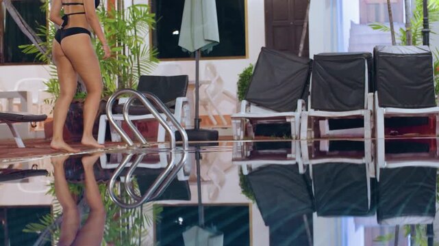Island resort poolside woman walking ladder, wearing bikini, stepping into pool with clear reflection, lounge chairs and potted palms lining tile deck, sunlit interior doorway in background, relaxed