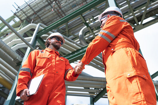 Two industrial engineers wearing orange safety uniforms and helmets shaking hands in refinery construction site, one holding documents, captured from low angle view with industrial pipes background.