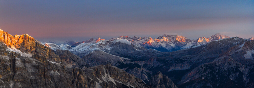 Aerial view of the rugged Alps mountains, snow-dusted peaks catch the warm glow of the rising sun, contrasting sharply with the cool shadows nestled in the valleys, Veneto, Italy.