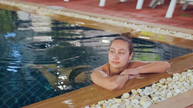 Woman relaxing at island pool edge, traveler resting chin on arms, gentle water ripples, tiled pool basin, pebble border, red deck chairs, reflective surface, evening glow, tranquil vacation mood