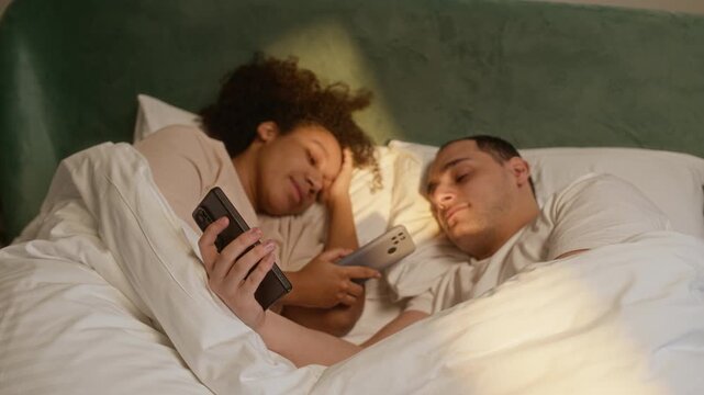 Medium shot of biracial married couple lying in bed, using smartphone, checking their social networks
