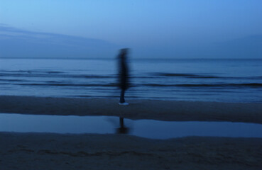 A blurred figure stands on a wet beach by the ocean under a twilight sky. The figure's reflection is visible in a puddle on the sand. © Robert
