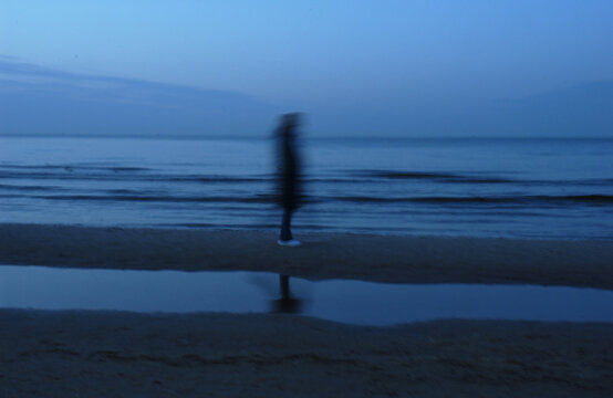 A blurred figure stands on a wet beach by the ocean under a twilight sky. The figure's reflection is visible in a puddle on the sand.