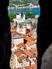 Scenic view of Kotor old town with red tiled roofs and church domes, framed by stone wall opening, overlooking bay, Mediterranean architecture, travel destination and calm summer atmosphere © Tishina