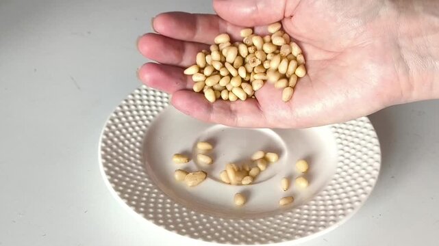 A handful of pine nuts in his hand. A woman's hand pours a kernel of cedar nuts into a white plate on the table
