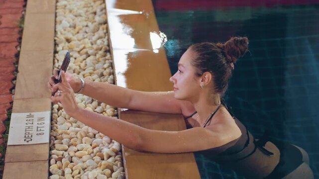 Pool selfie at island resort, woman leaning on pool edge holding phone, illuminated stones and warm lights reflecting on water, candid smile and relaxed pose capturing nighttime memory for social