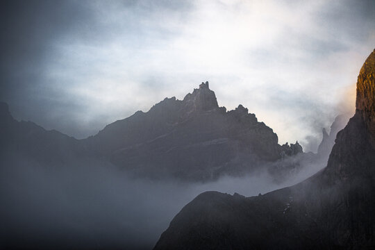 Aerial view of jagged peaks pierce through the swirling mist, a symphony of grey stone against the soft, diffused light, Val Badia, Trentino-Alto Adige, Italy.