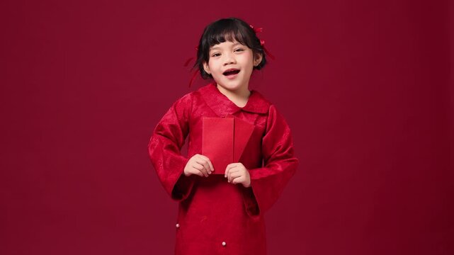 The cheerful Asian child smiles brightly, holding a red envelope and saying New Year's greetings in Chinese. Her bright red dress reflects the joyful festive atmosphere.