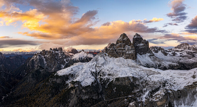Aerial view of rugged, snow-dusted peaks pierce the skyline, bathed in the warm glow of a setting sun, Tre Cime di Lavaredo, Veneto, Italy.