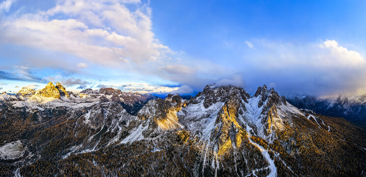 Aerial view of Tre Cime di Lavaredo and Cadini of Misurina in golden sunlight against a backdrop of azure skies and swirling clouds, Veneto, Italy.