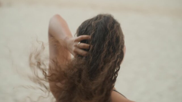 Multiracial woman on island beach back view hair flip into wind, flowing curly strands, sandy shoreline backdrop, casual swimwear, relaxed movement, playful traveler and local islander vibes,