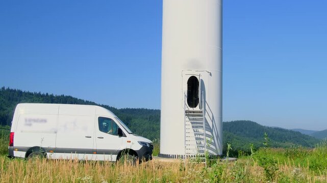 Service van parked near windmill support with open technical entrance in green field slow motion. Brigade checks condition of wind turbine in valley