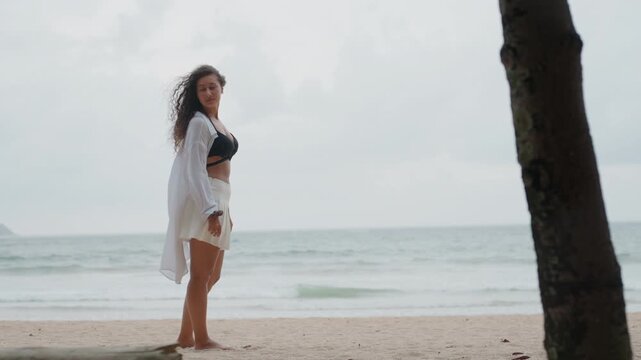 Full body woman standing on island shore with ocean waves, palm trunk foreground, overcast sky, flowing white wrap and swimsuit, traveler pausing in contemplative gaze, moody tropical escape